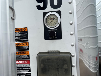 Trailer rear showing a circular gauge labeled 'PULL TO DUMP' with a metal plate and stacked warning stickers; part of a Timpte hopper trailer.