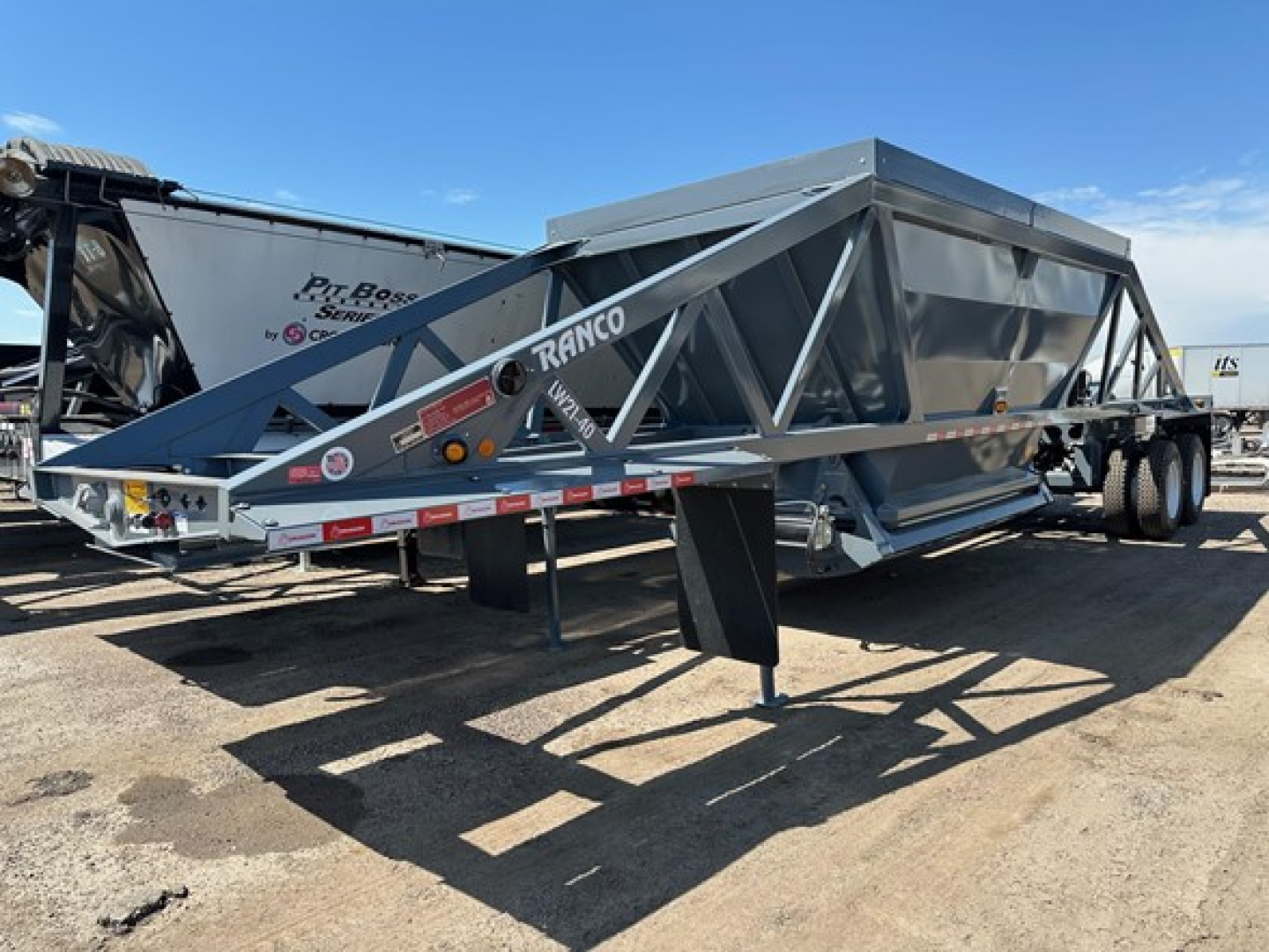 Lw21-40 Ranco dump trailer in a yard, gray tri-axle frame with lattice truss sides, showing 'RANCO' branding on beam and ramp legs, tires and hitch visible.
