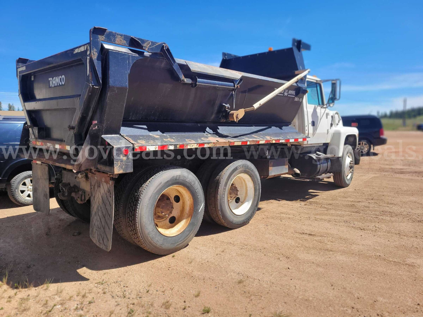 Dump truck rear and side view showing large black dump bed, white cab, dual rear axles with tan wheels, and hydraulic piston. Visible text on dump bed: 'DUMP' and 'E T H O' likely. Brand insignia and generic truck components visible.