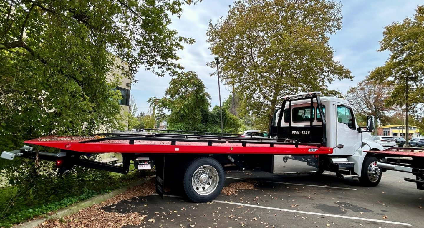 Kenworth T280 tow truck with a long red flatbed and white cab, parked among trees in a lot.