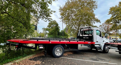 Kenworth T280 tow truck with a long red flatbed and white cab, parked among trees in a lot.