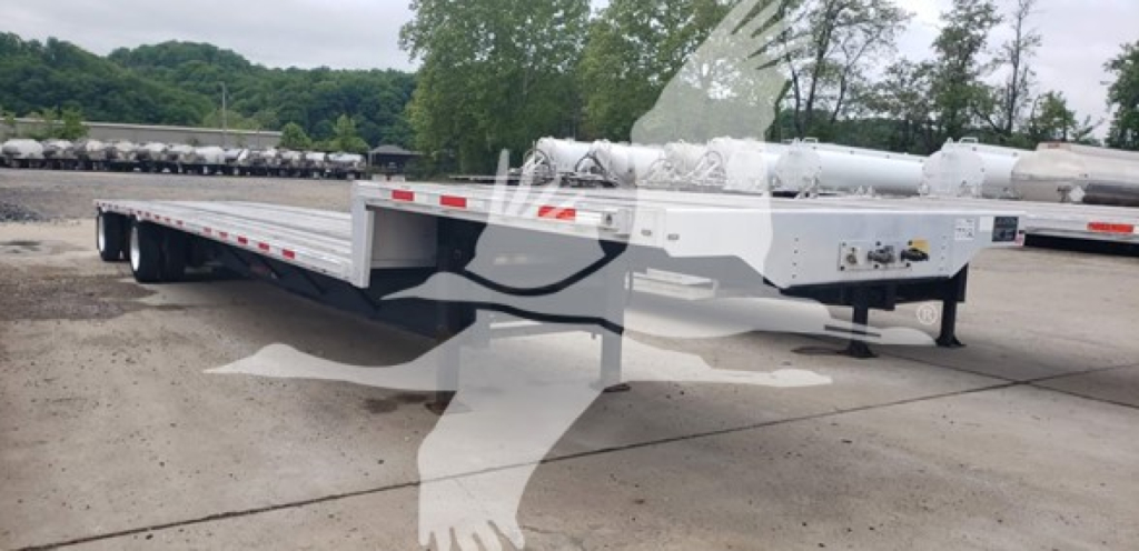 Flatbed trailer in a yard, side view showing long deck and drop deck features; no legible serials or labels visible on the trailer body.