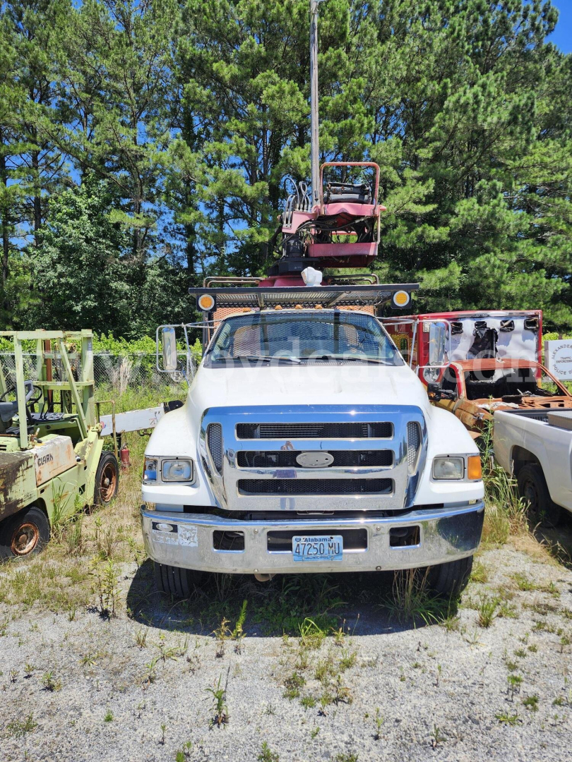 Front view of a white Ford F-750 XL truck with a grapple hoist on top, chrome front bumper, license plate visible, and other equipment in the background.