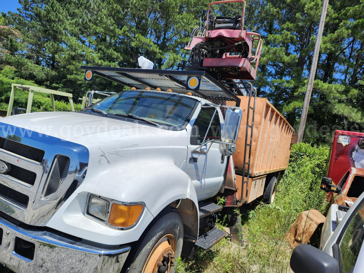 Ford F750XL grapple truck showing front cab and flatbed with trailer and equipment mounted, white cab with blue fender, orange dumpster bed, equipment on top stake, outdoor yard.
