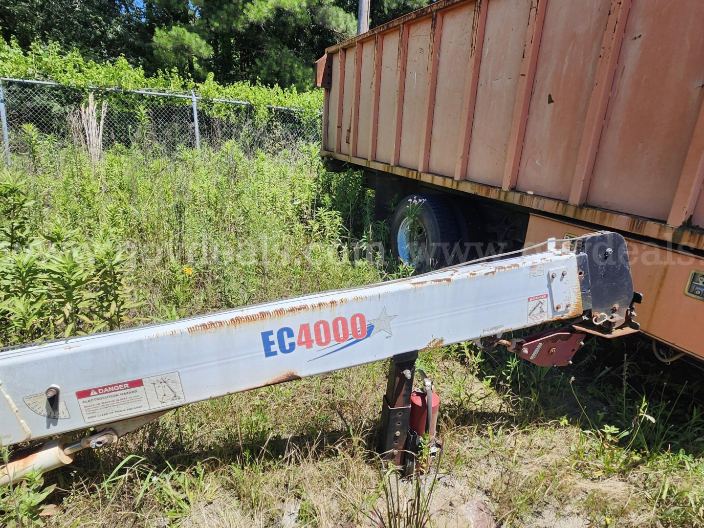 Ford F750XL grapple truck in-field showing EC4000X crane arm and trailer hitch with overgrown grass and a rust-colored container.