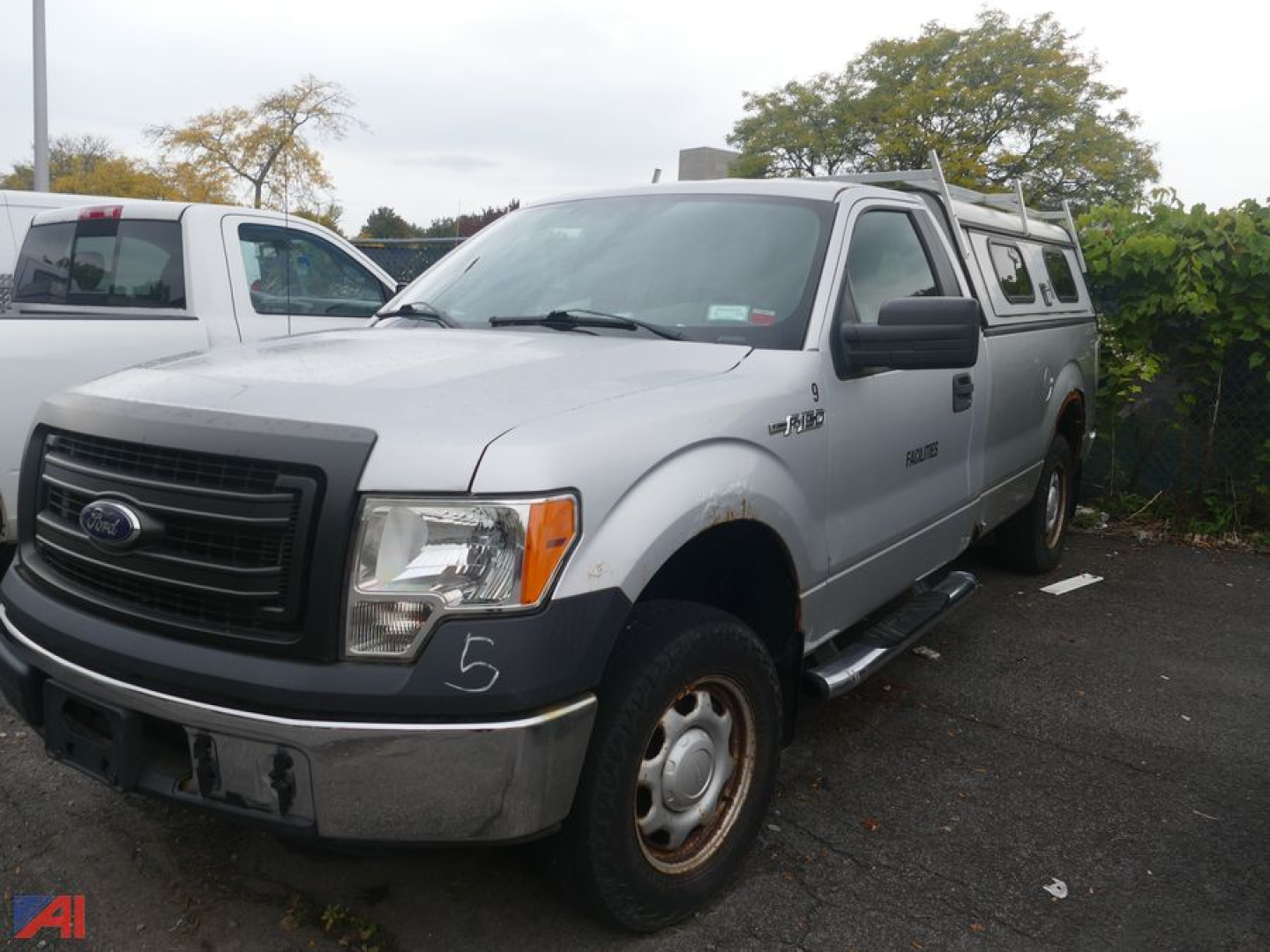 Ford F-150 pickup truck with camper shell, silver body, front grille, headlights, and running boards in a parking lot.