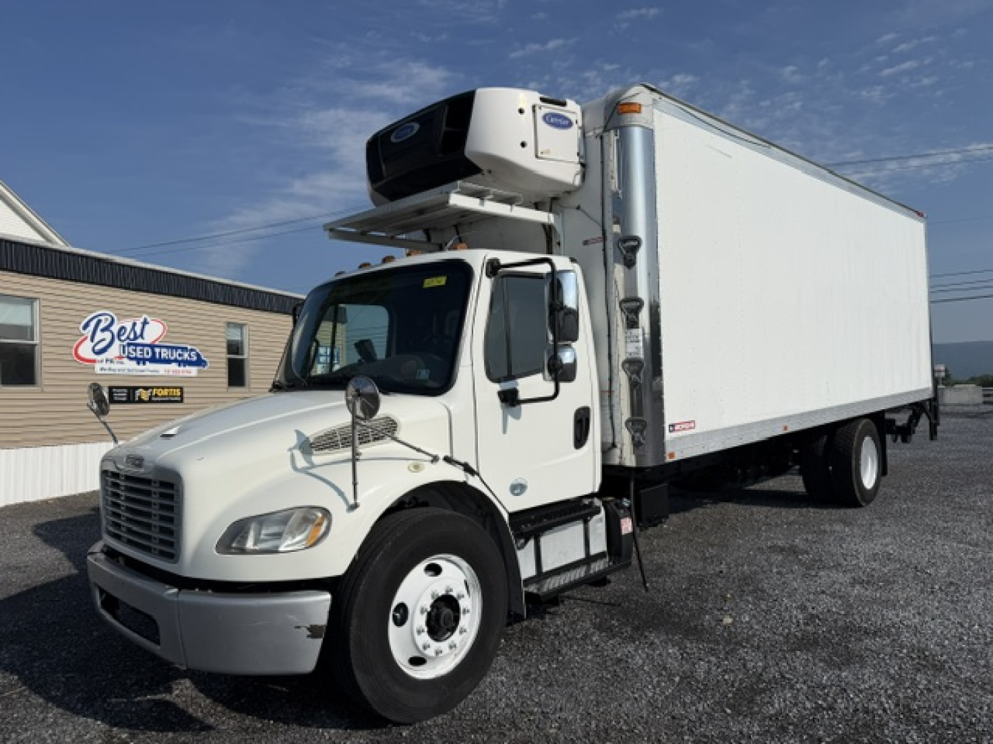 Freightliner M2-106 refrigerated truck with a mounted APU unit above cab and large box body in a parking lot.