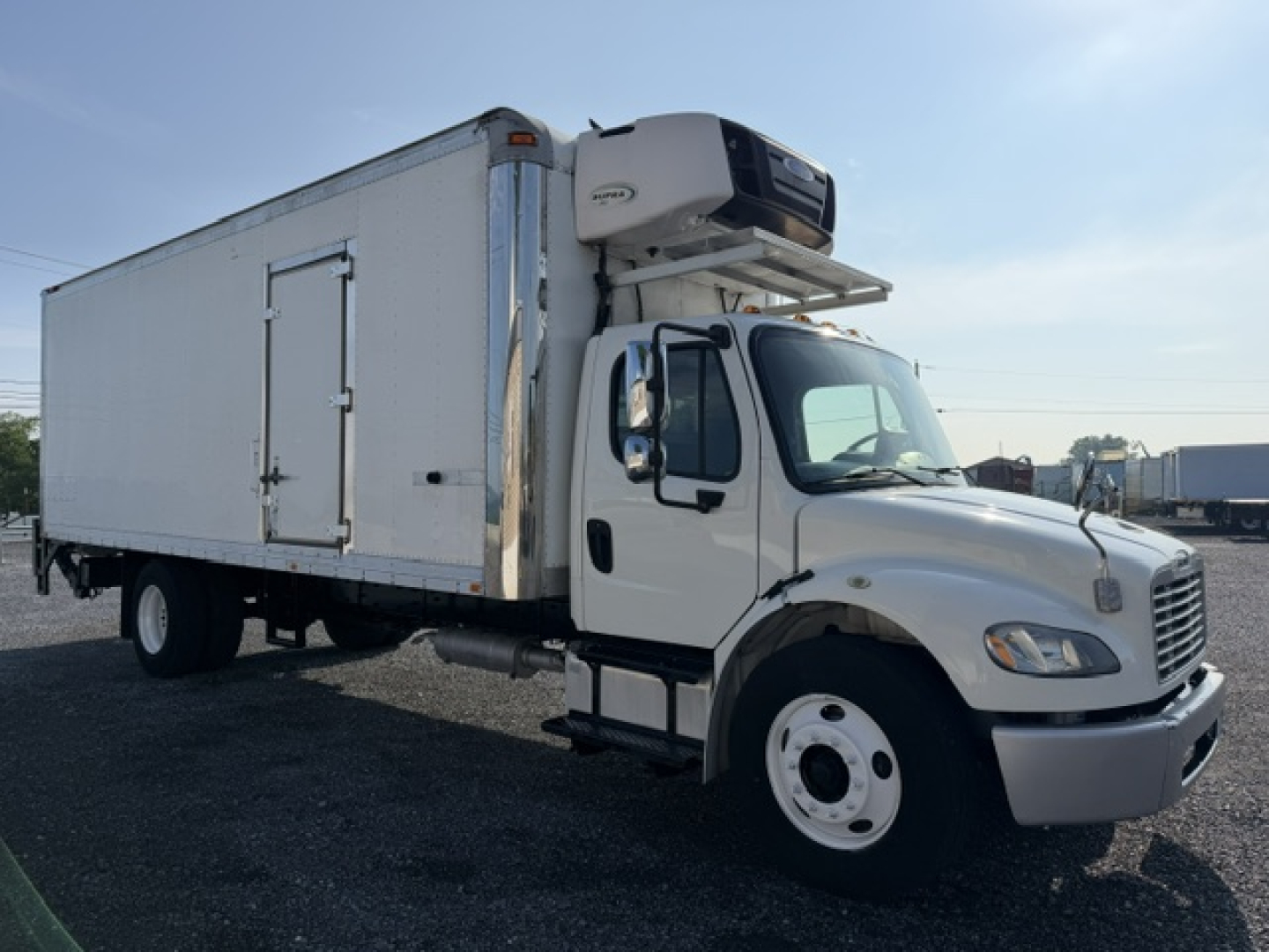 Freightliner M2-106 refrigerated truck with a mounted refrigeration unit on top of the cargo box, front cab visible, white exterior, side door on box.