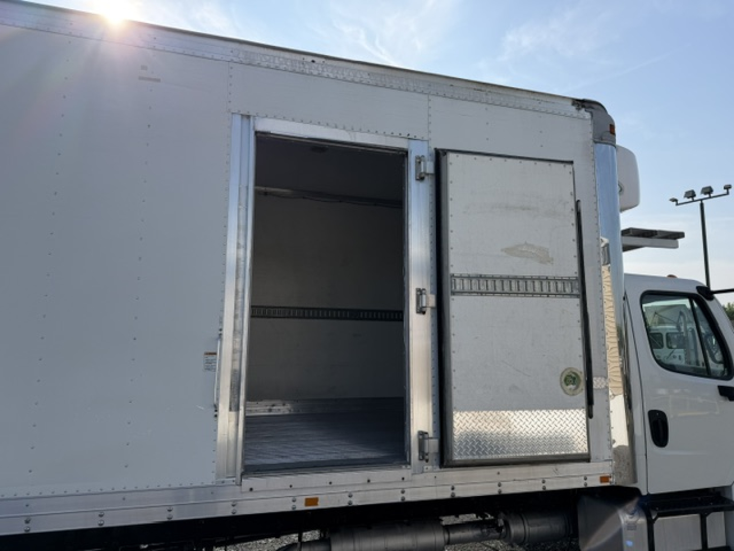 Freightliner M2-106 refrigerated truck side door open showing empty aluminum cargo box with metal floor and hinge joints.