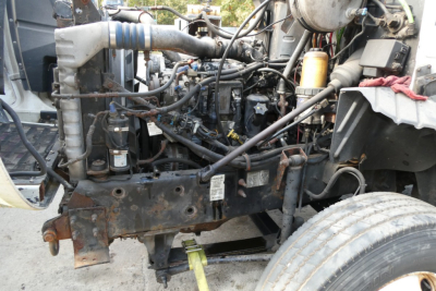 Truck engine bay of a 2006 Mack CXN613 Conventional Sleeper showing exposed engine components, hoses, and frame; visible front suspension and wheel.