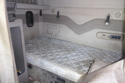 Truck sleeper cab interior with a single metal quilted mattress, side storage pocket, and control panel on the wall; light gray fabric panels and curved decorative trim.