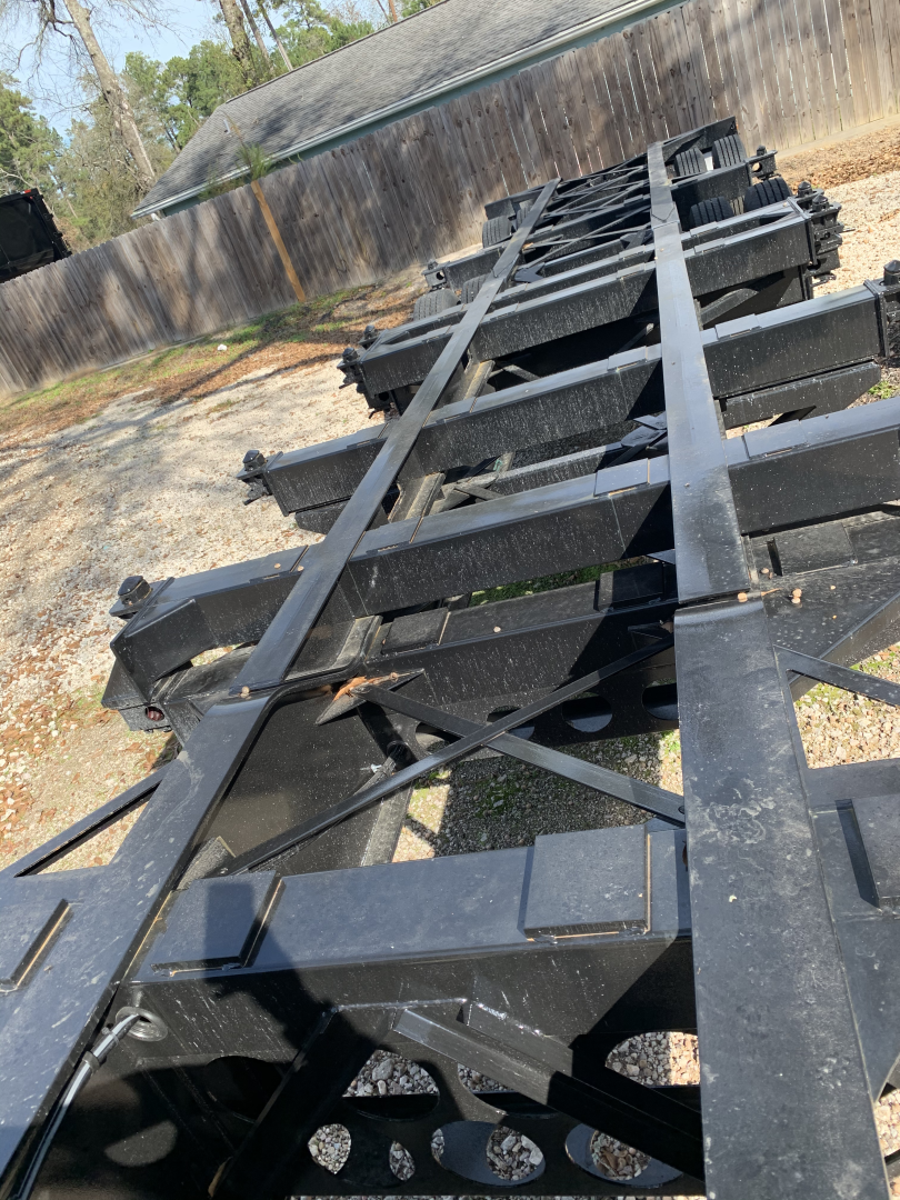 Trailer chassis frame showing steel I-beams and cross members with some hydraulic components and wheels in the distance.