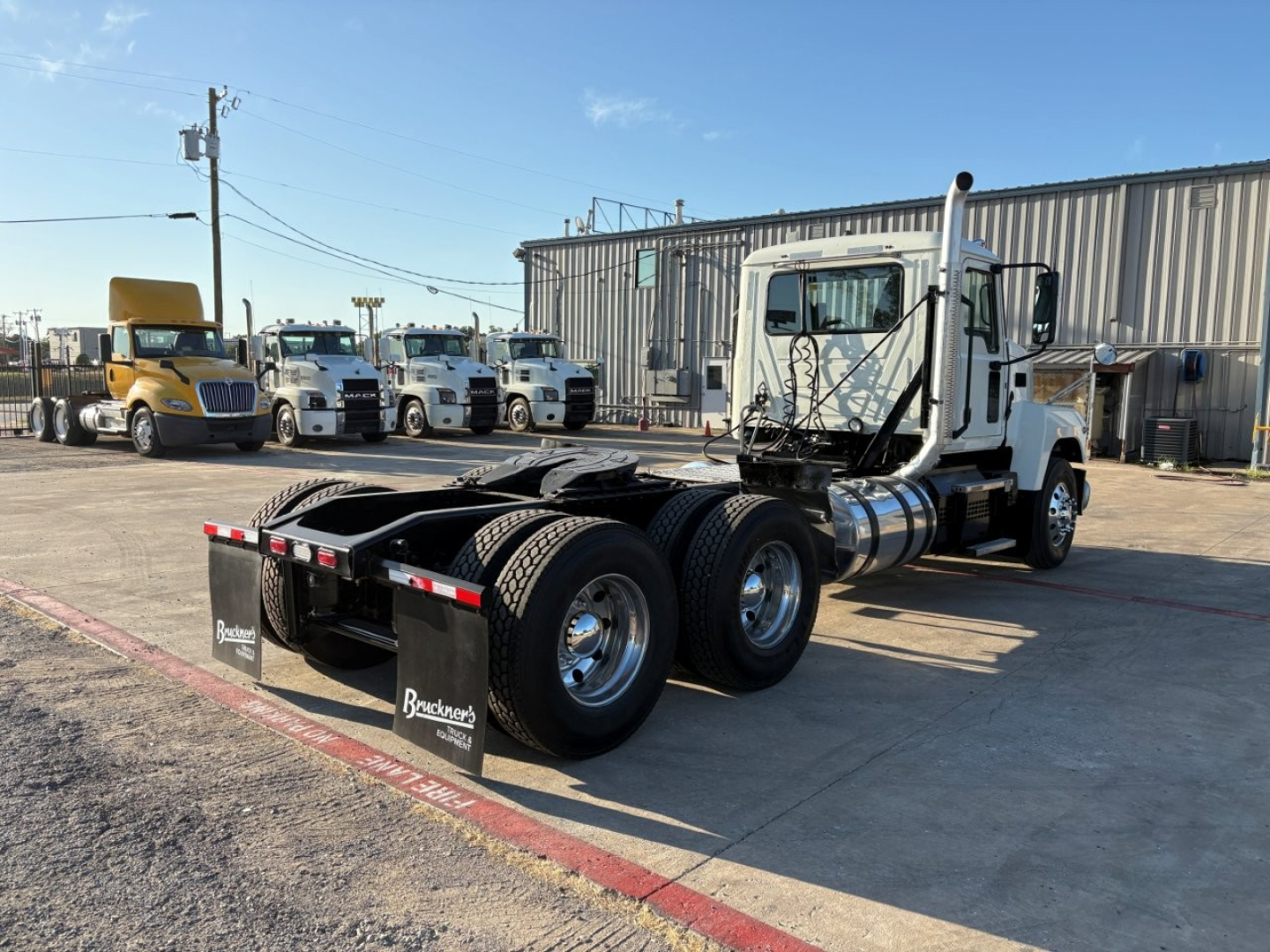 2020 Mack PI64T tractor rear view with fifth wheel and dual rear axle in a lot; text on mudflaps reads ‘Bradsen’ or similar; some trucks and a building in background.