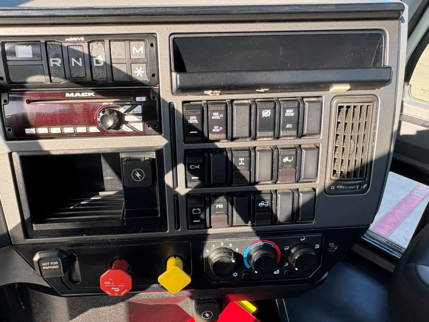 Mack PI64T dashboard cockpit with gear selector showing R N D M, red Mack badge, row of switches, climate controls, and three colored knobs (red, yellow).