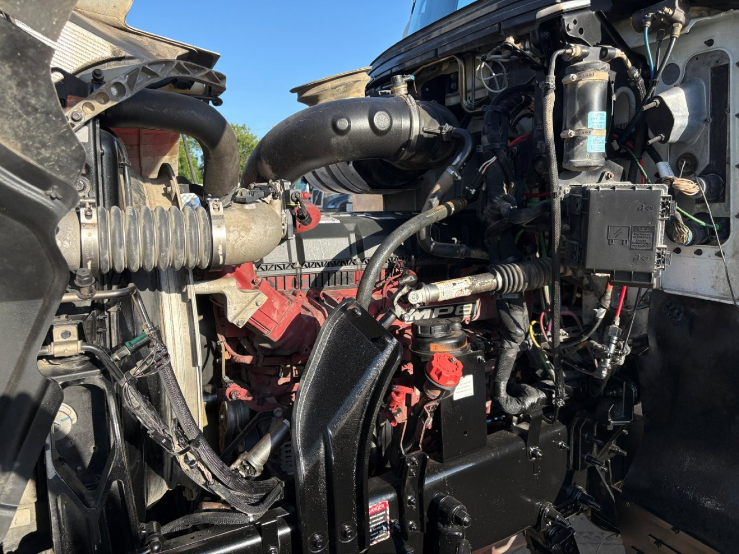 Engine bay of a 2020 Mack PI64T conventional daycab showing the engine with hoses, battery box, and wiring harnesses.