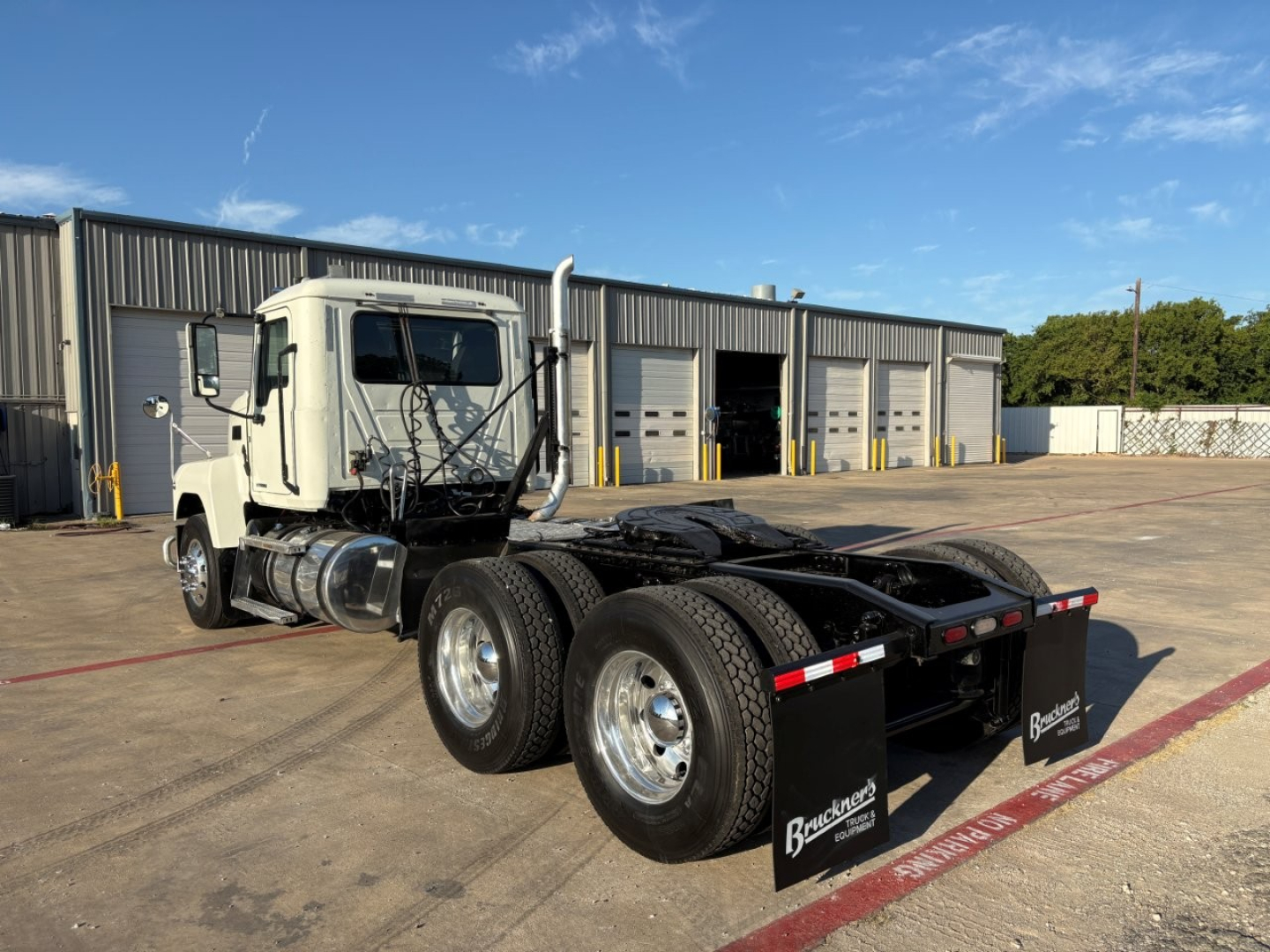 Tractor unit of a 2020 Mack PI64T with three rear axles, white cab, chassis and dual rear tires, fifth wheel visible, in a yard with service bays.