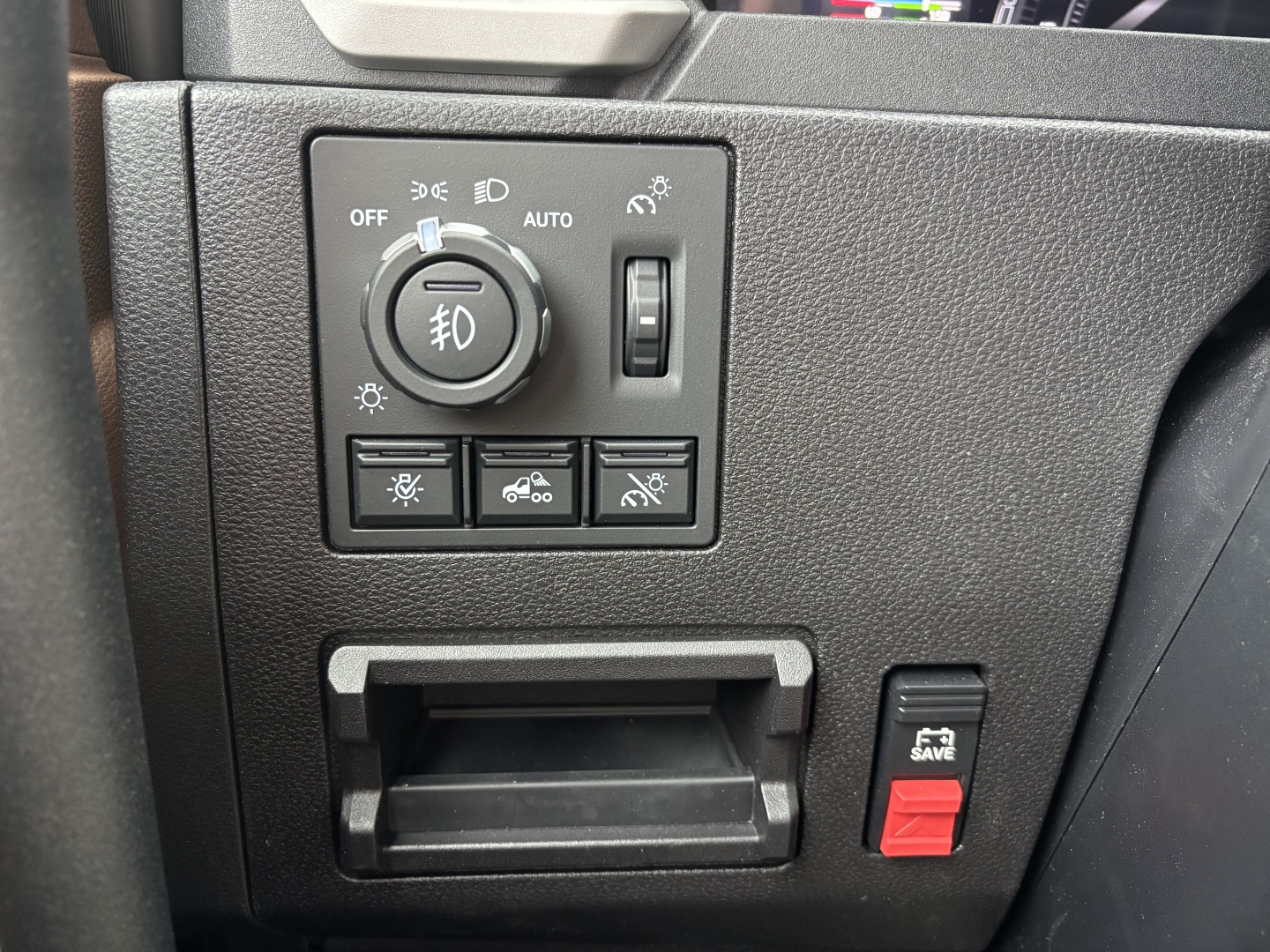 Truck dashboard control panel with headlight switch in center and several buttons below; small cubby/ashtray area and a red toggle switch labeled 'SAVE' on the right side.