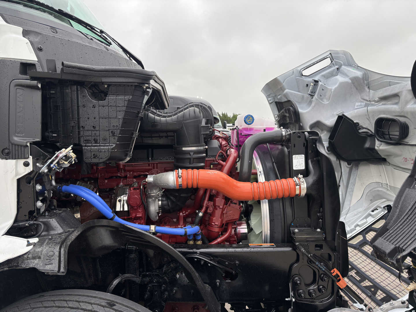 Mack PR64T76M engine bay, with red engine block, blue and orange hoses, and front cab components visible.