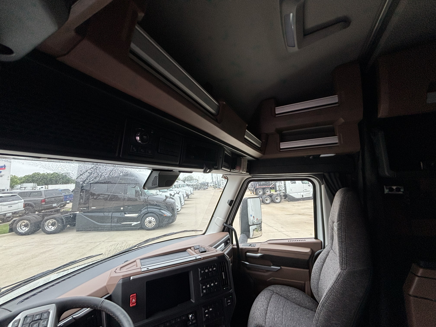 Interior of 2027 Mack PR64T76M cab showing driver seat, dashboard, overhead storage, and windshield view of trucks lot.