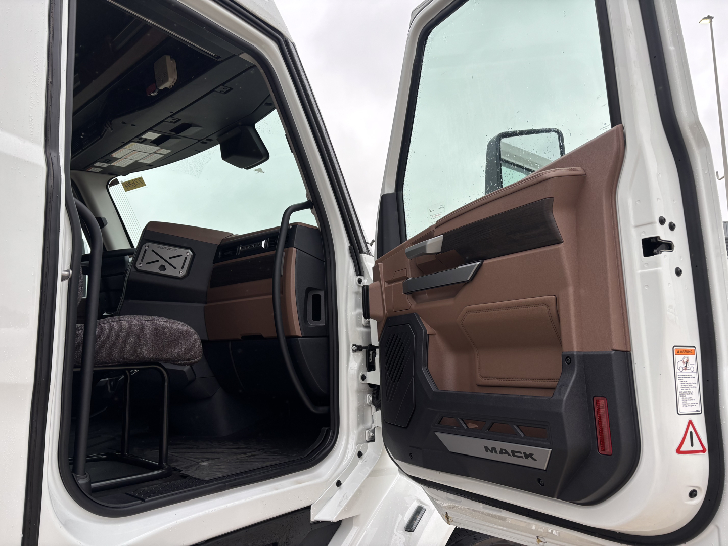 Cab interior of a white Mack truck with tan and black door panels; 'MACK' branding on door sill; partial dashboard; open driver door.
