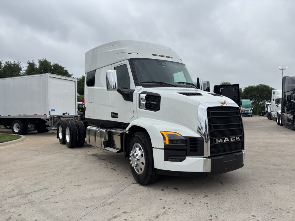 New 2027 Mack PR64T76M conventional sleeper truck shown in a dealership lot, front three-quarter view, white exterior, 6x4 tractor with dual rear axles, Mack emblem on grille.