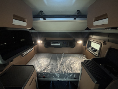Interior sleeper cab of 2027 Mack PR64T76M showing bed with plastic cover, overhead cabinets, side windows, and control panel mounting in center.