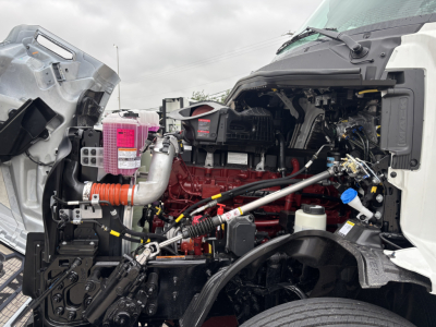 Tractor truck engine bay view showing red engine block with various hoses and metal brackets; pink relay box and air intake housing visible; exposed wiring and components in the engine bay.