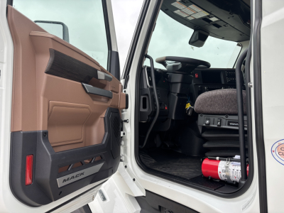 Truck cab interior door open showing Mack PR64T76M model; front interior with steering wheel, driver seat, dashboard; red fire extinguisher visible on floor near door.