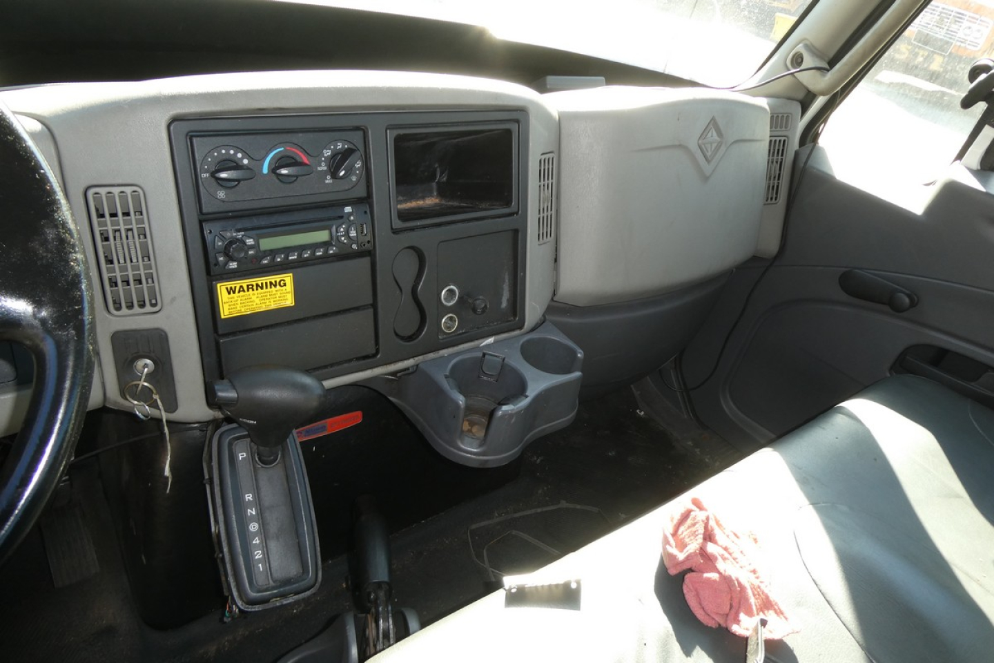 Interior of a truck cab with dashboard controls, center stack, gear shifter, cup holders, and a small screen area. Visible labels include a yellow warning sticker and various dials.