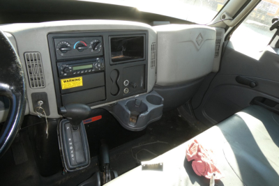 Interior of a truck cab with dashboard controls, center stack, gear shifter, cup holders, and a small screen area. Visible labels include a yellow warning sticker and various dials.