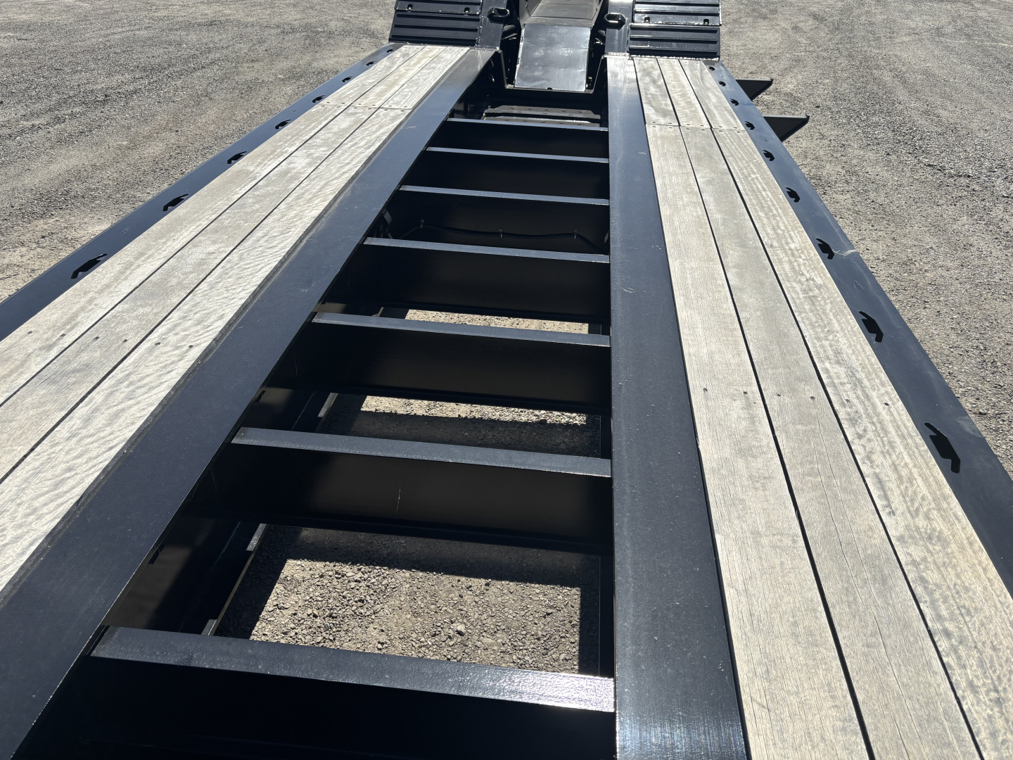 Lowboy trailer bed showing steel frame with wooden deck planks, center ladder-like support, and side rails; top view toward rear with wheel wells not visible.