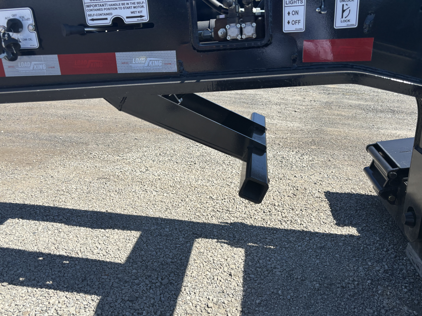 Trailer hitch area of a Load King lowboy trailer showing a support leg and hydraulic mechanism with warning labels and reflective tape.
