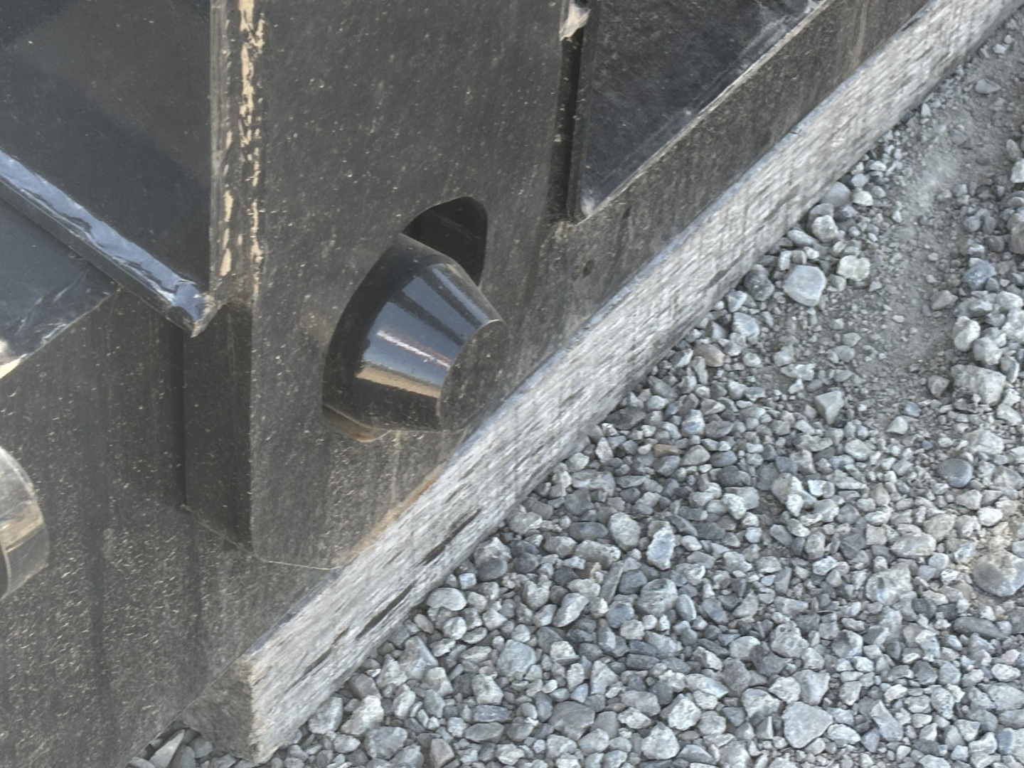 Load King trailer riser or wheel area showing black painted metal with a cutout and a shiny conical pin, gravel ground visible