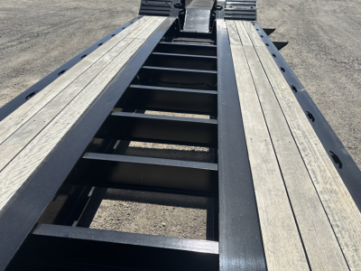 Lowboy trailer bed showing steel frame with wooden deck planks, center ladder-like support, and side rails; top view toward rear with wheel wells not visible.