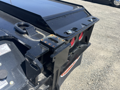 Trailer rear corner showing black steel frame with red taillights and a tapered hitch area; visible labels and bolts, metal brackets, and a small white sticker.