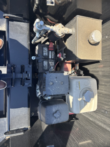 Machine component close-up: engine-like module with hoses, metal fittings, and a rectangular reservoir on a trailer chassis.