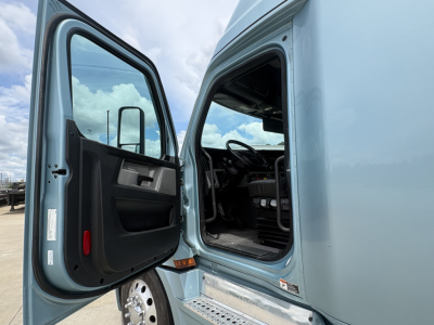 Truck cab interior view with door open showing Freightliner Cascadia 126 sleeper cab dash and steering wheel area.
