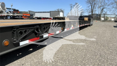 Lowboy trailer side view showing steel frame, black components, white and red reflective tape, and wooden deck with wheel axles visible.