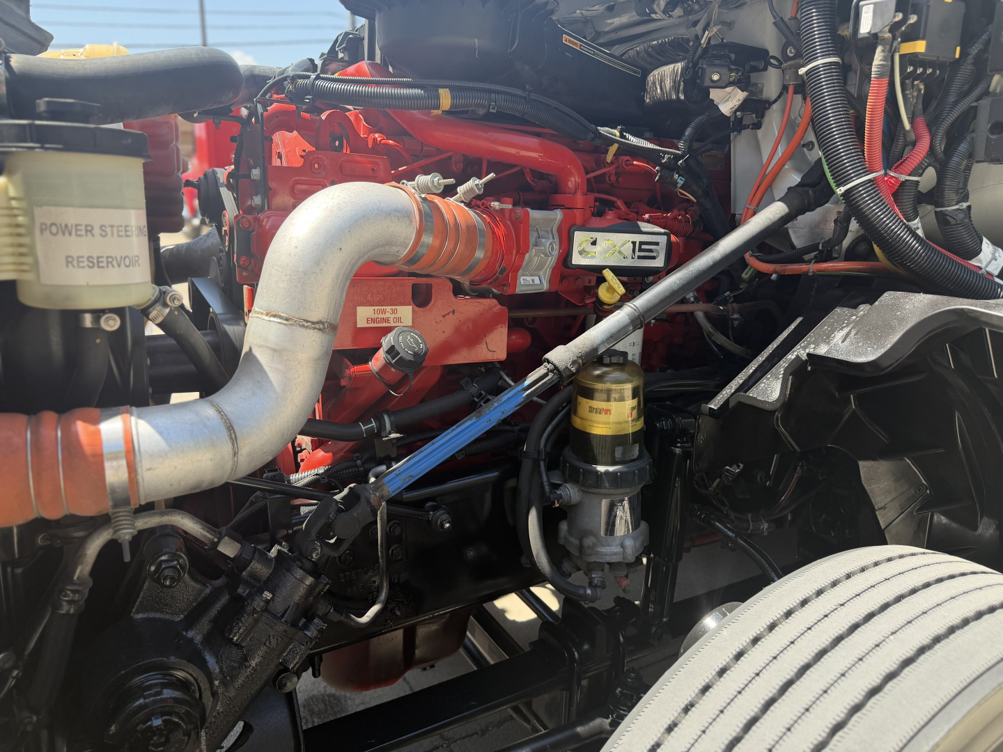 Truck engine bay showing large metal intake pipe, coolant reservoir, power steering reservoir, and yellow engine badge in red engine area.