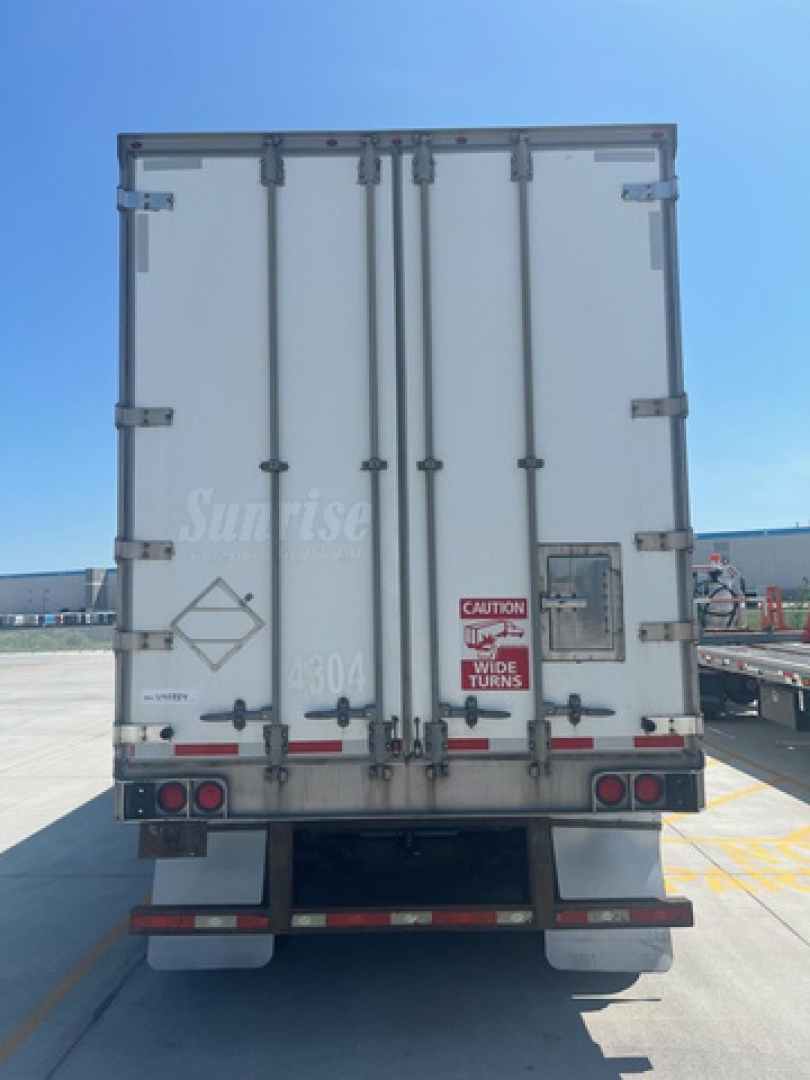 Rear of a white utility van/trailer with double doors, CAUTION wide turns sticker, taillights, and rear bumper, Sunrises logo faintly visible.