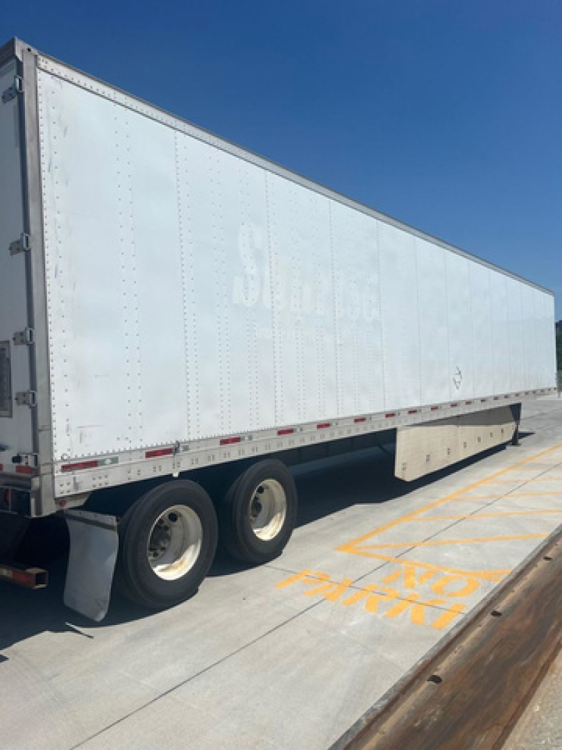 Trailer side view showing a large white utility van trailer with dual rear axles and faint logo near the top.