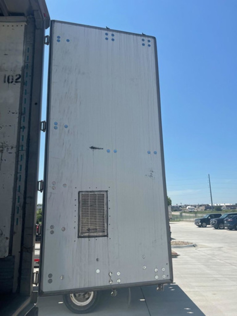 Trailer side panel of a utility van with ventilation grate and riveted aluminum skin; visible wheels and open trailer door edge.