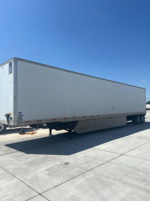 Long white utility trailer in a paved lot, side view with landing gear, no visible branding except small numbers near the front. Clear blue sky.
