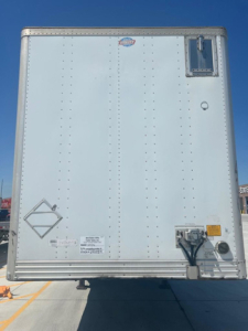 Rear view of a white 2012 utility van trailer with riveted panels, small square access door upper right, bumper guard bottom center, and various decals near lower left.