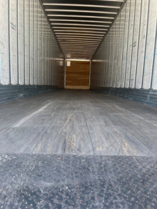 Interior of a trailer showing empty cargo space with wooden bulkhead at the far end. Rust-free steel walls with corrugated side panels and a diamond plate floor.