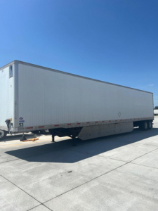 Long white utility trailer in a paved lot, side view with landing gear, no visible branding except small numbers near the front. Clear blue sky.