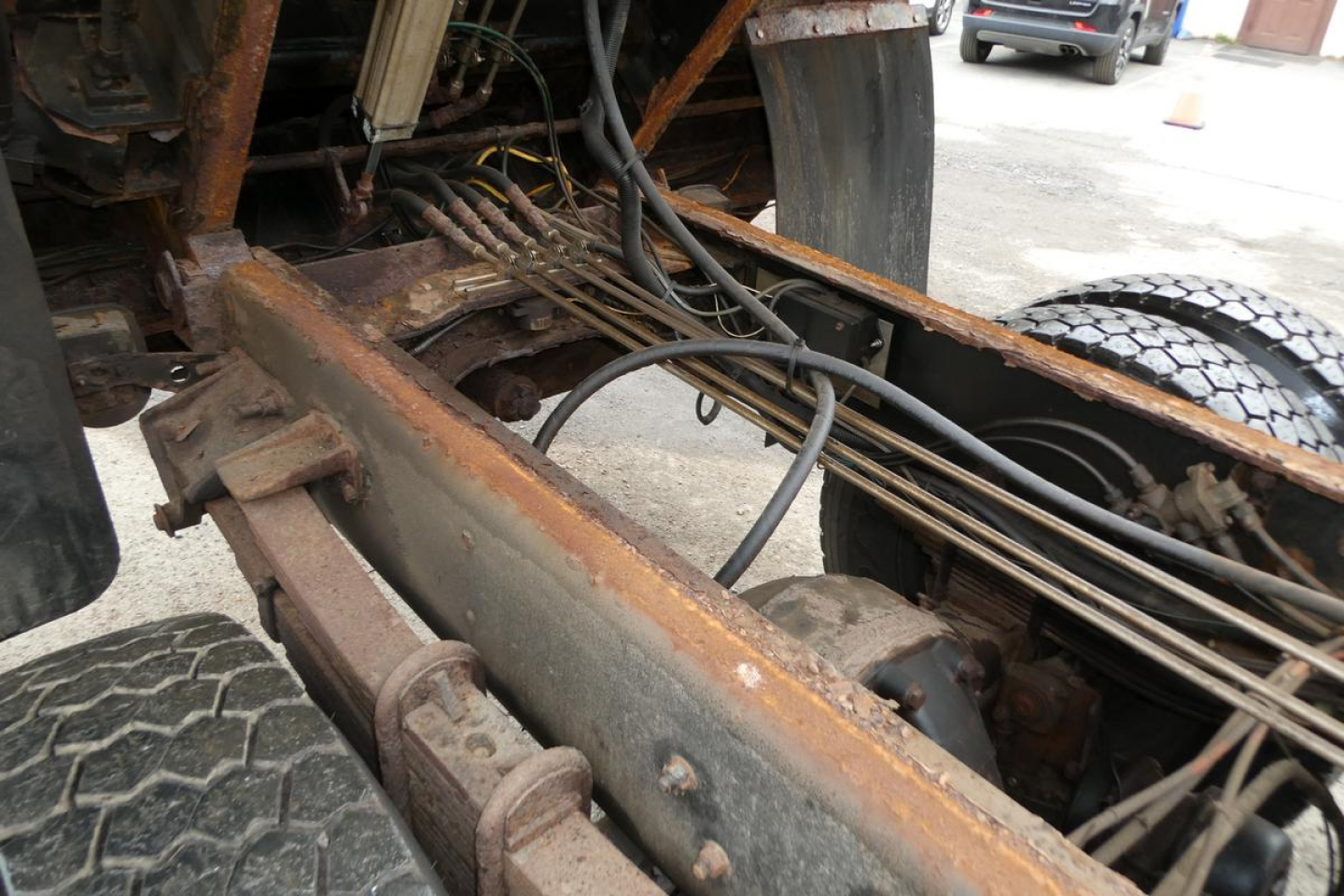 Truck chassis underside showing frame rails, leaf springs, and drive components with rusted metal surfaces and wiring running along the frame.