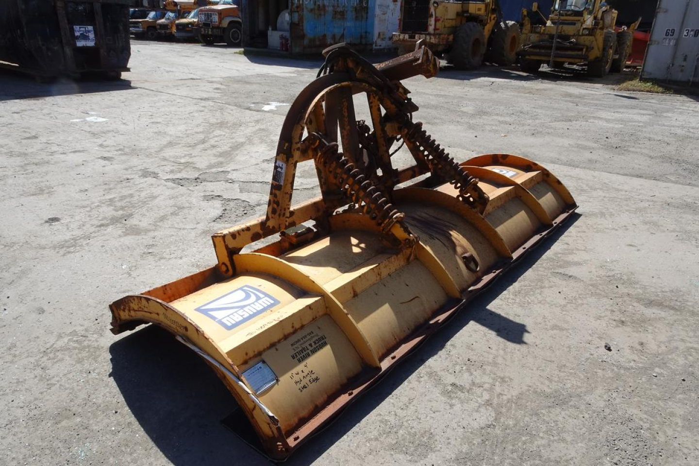 Heavy-duty Mack Granite GU712 bucket attachment with hydraulic linkage and wear plates, yellow metal, worn stickers, in outdoor yard.