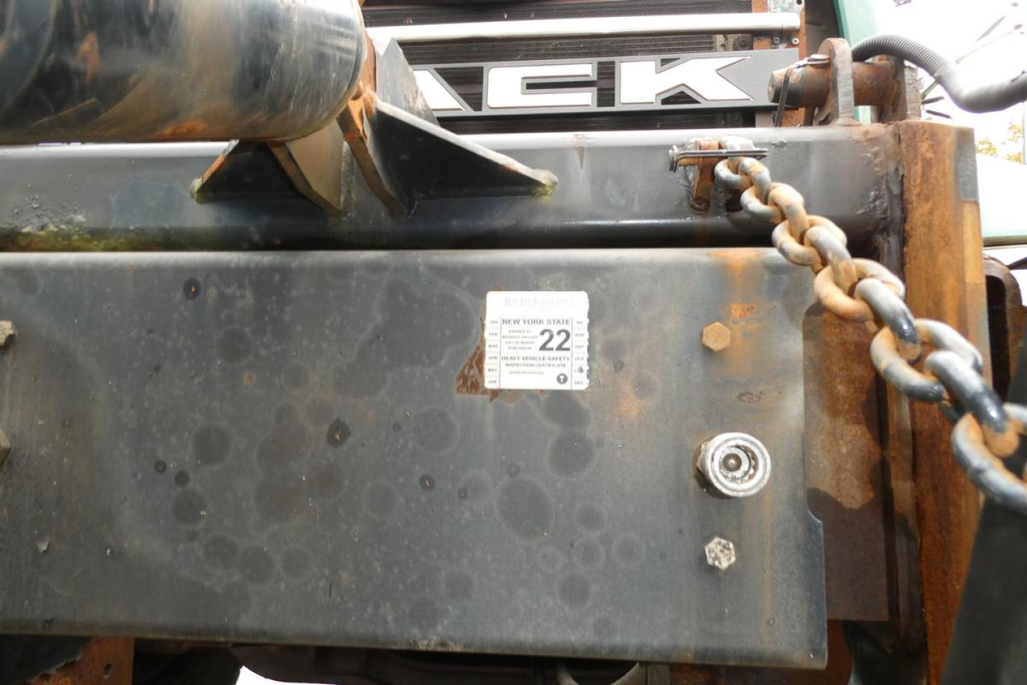 Truck front end showing Mack logo and attached chain with rust on metal panel; small white sticker with numbers; hydraulic hose visible.