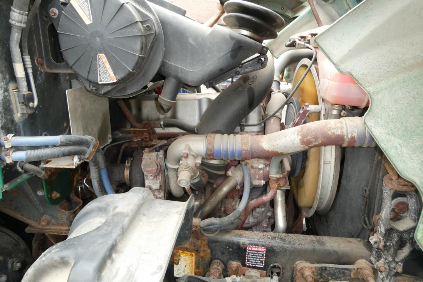 Mack GU Granite engine bay showing coolant reservoir, hoses, belts, and various hydraulic lines in a used dump truck.